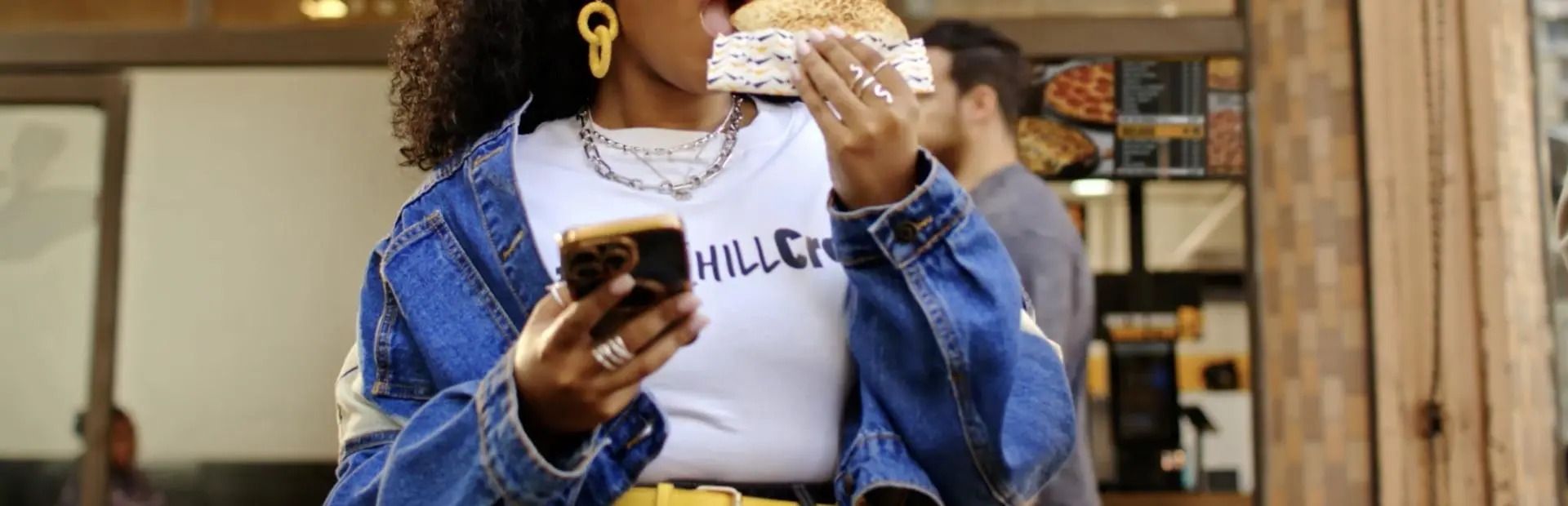 A woman wearing a denim jacket and yellow earrings holding a smartphone while eating a sandwich.