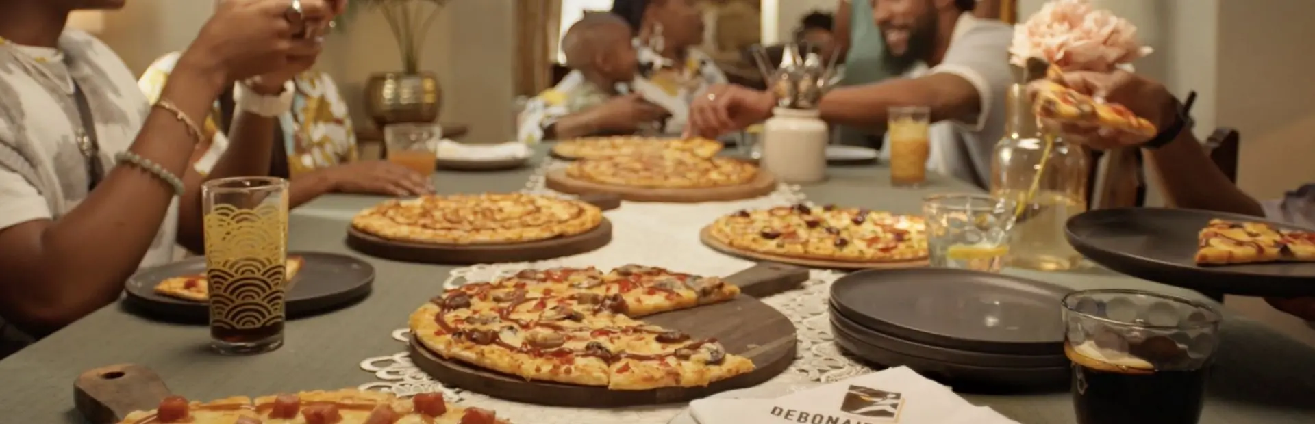 Family and friends gathered around a dining table sharing various freshly baked pizzas and drinks.