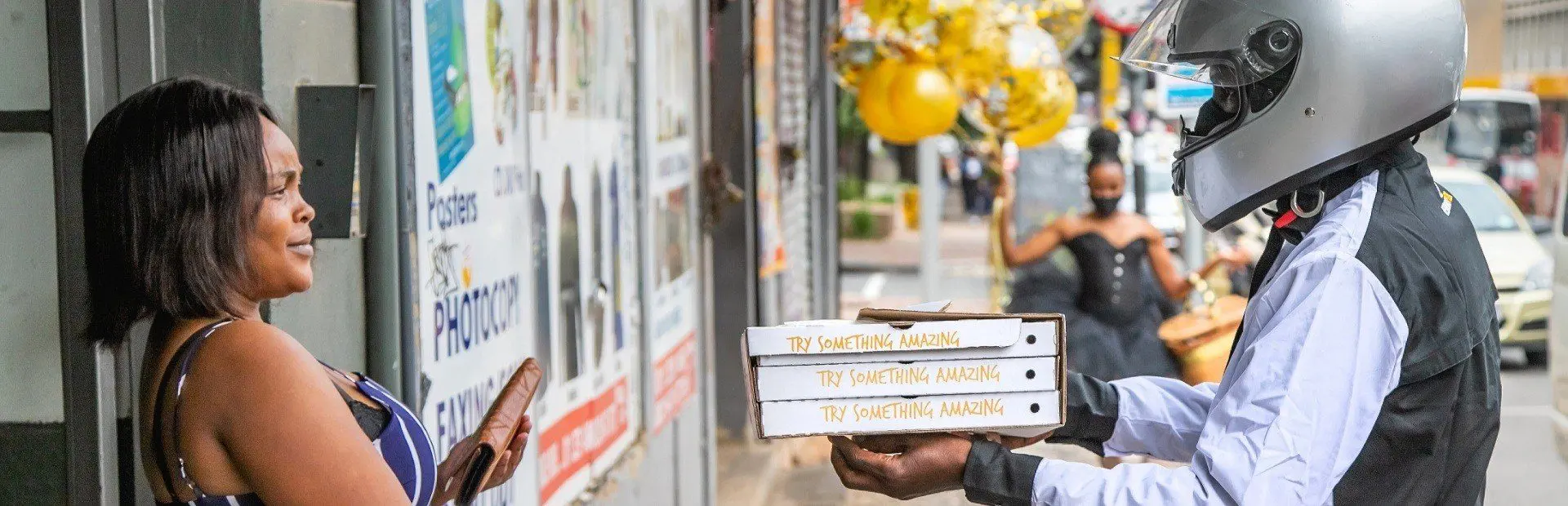 Delivery driver wearing a helmet hands three pizza boxes labeled Try Something Amazing to a woman on a city sidewalk.